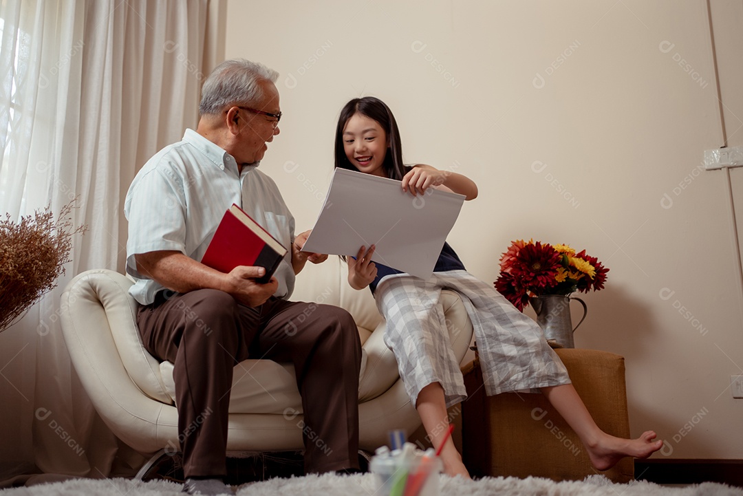 O vovô bonito asiático está segurando um livro enquanto sua neta está desenhando em um papel