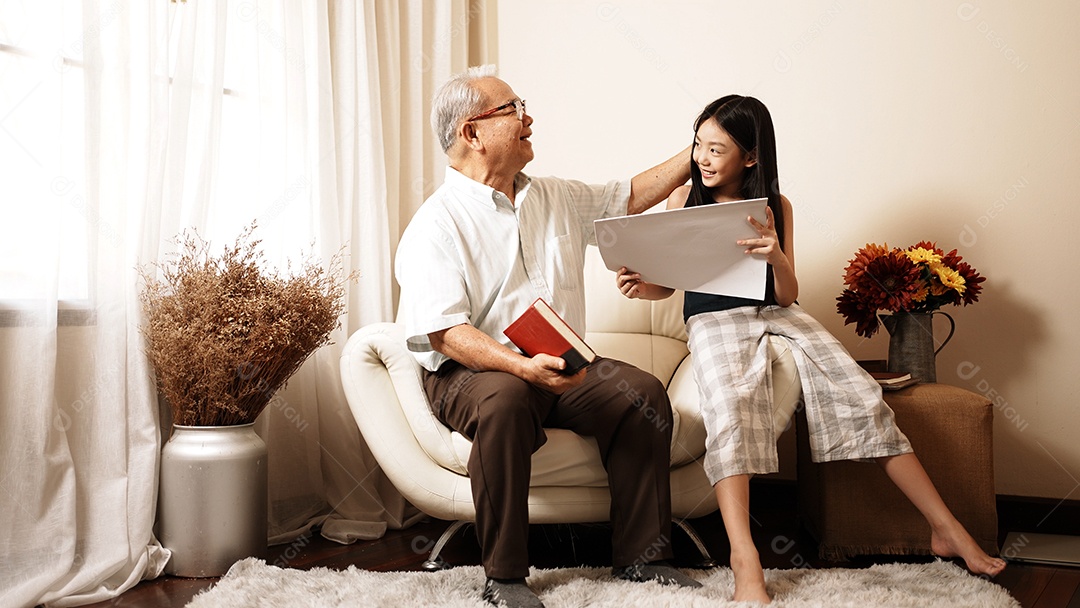 O vovô bonito asiático está segurando um livro enquanto sua neta está desenhando em um papel