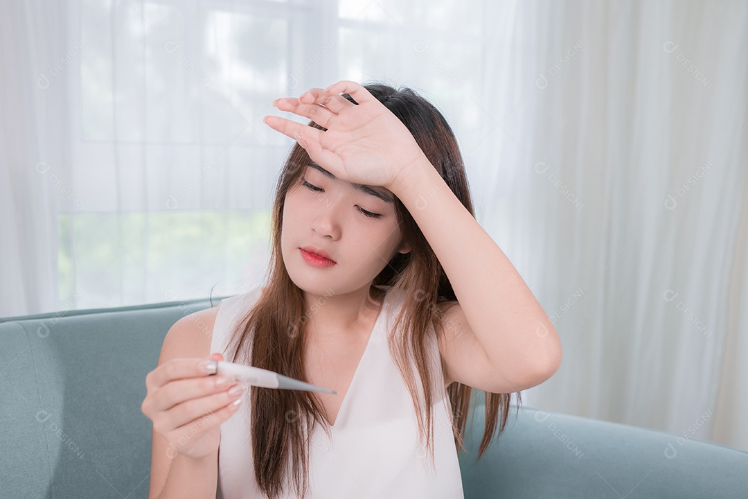 Mulher asiática verificando sua temperatura com um termômetro no sofá na sala de estar