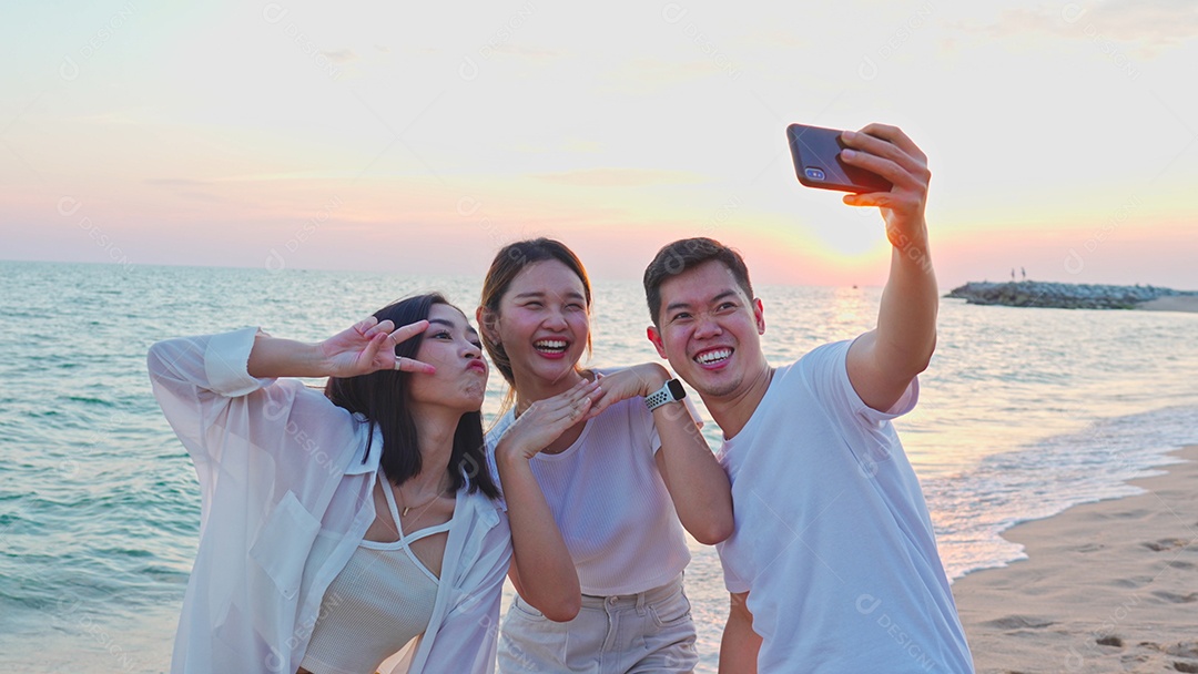 Grupo de amigos jovens adultos felizes tirando selfies no smartphone em uma praia ao pôr do sol