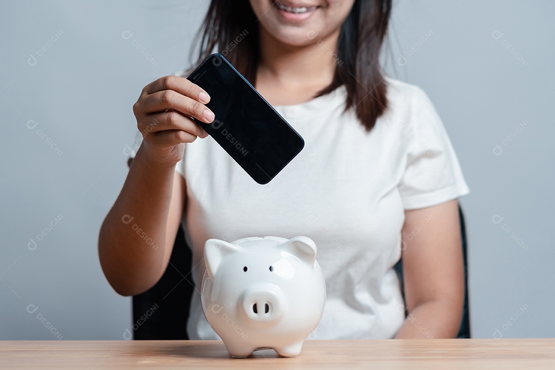 Mão de mulher segurando celular com tela preta e cofre de porquinho sobre a mesa