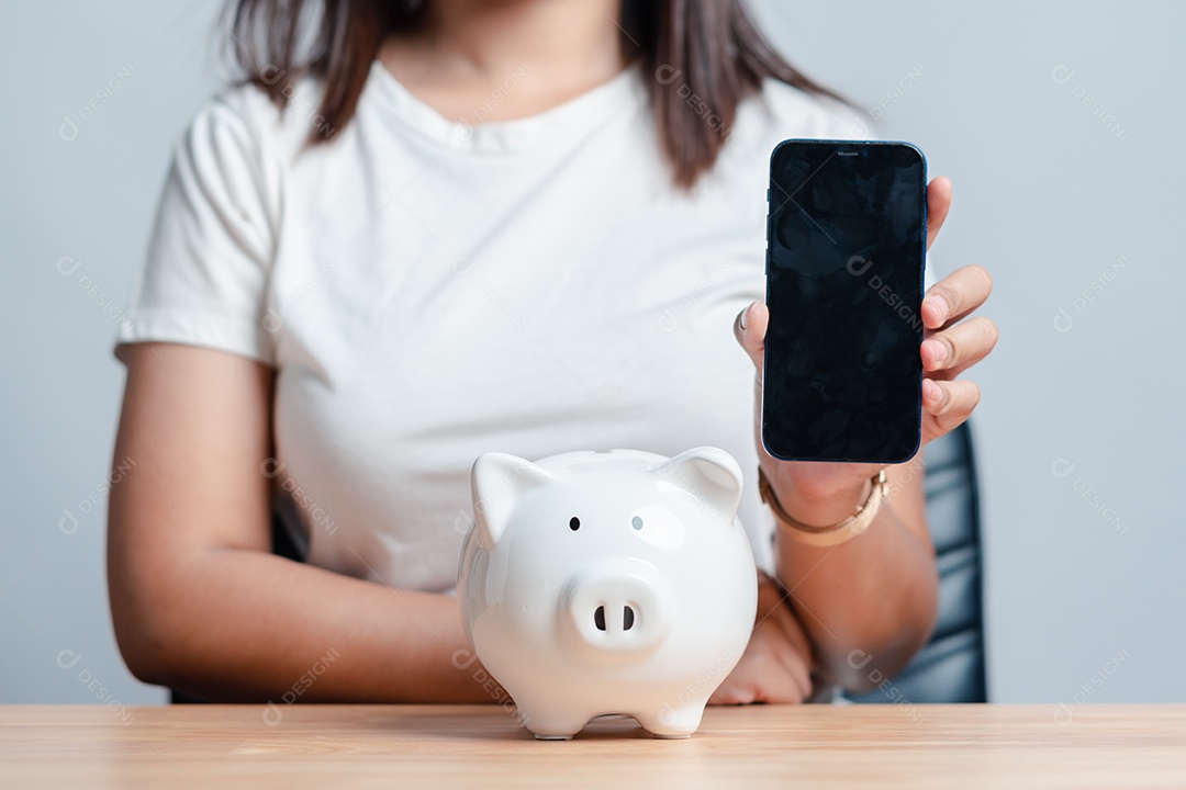 Mão de mulher segurando celular com tela preta e cofre de porquinho sobre a mesa