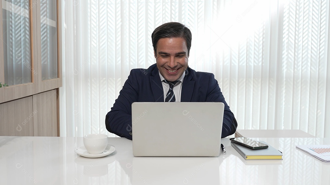 O empresário sorri feliz por receber uma boa notícia do laptop enquanto está sentado em uma mesa em um escritório.