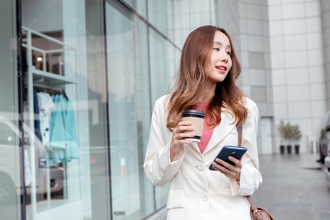 Linda empresária asiática usa smartphone segurando copo de papel com bebida quente