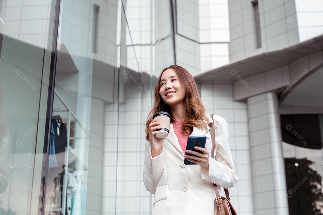 Linda empresária asiática usa smartphone segurando copo de papel com bebida quente