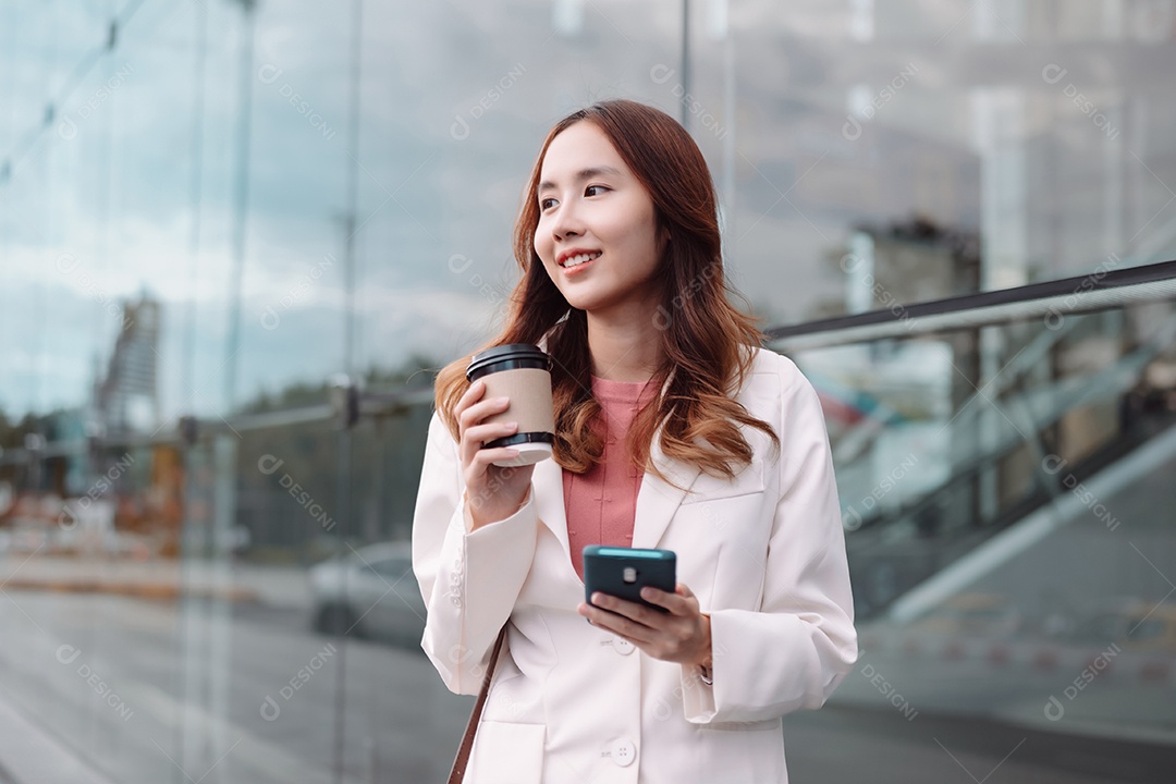 Linda empresária asiática usa smartphone segurando copo de papel com bebida quente
