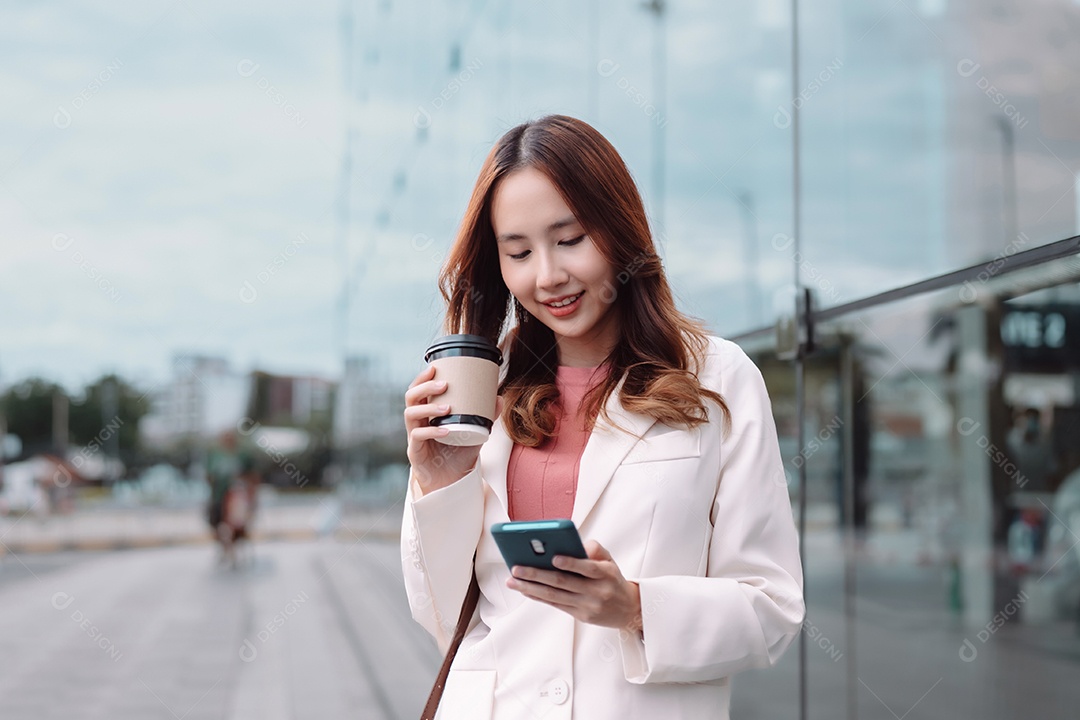 Linda empresária asiática usa smartphone segurando copo de papel com bebida quente