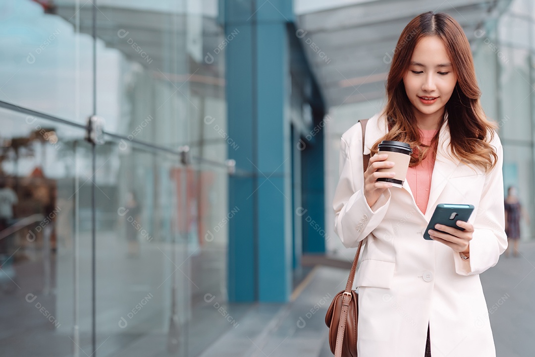 Linda empresária asiática usa smartphone segurando copo de papel com bebida quente