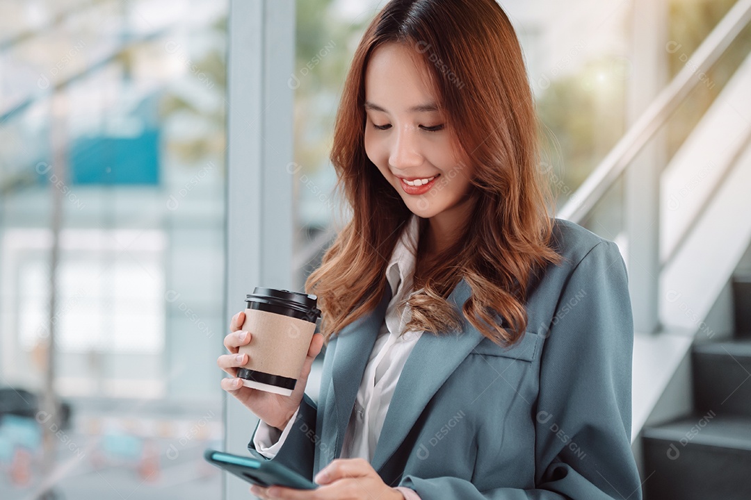 Linda empresária asiática usa smartphone segurando um copo de papel com bebida quente