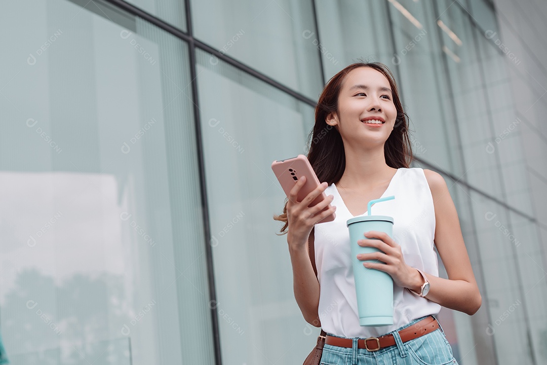 Empresária asiática sorridente usa smartphone e segura copo ecológico reutilizável