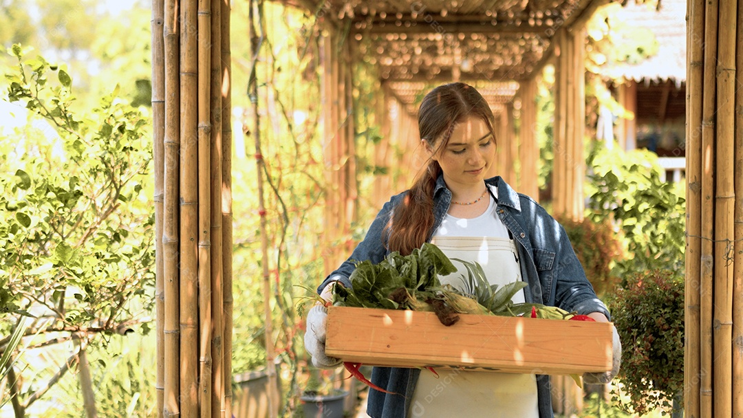 Mulher jovem feliz segurando caixote de madeira com legumes orgânicos frescos da fazenda