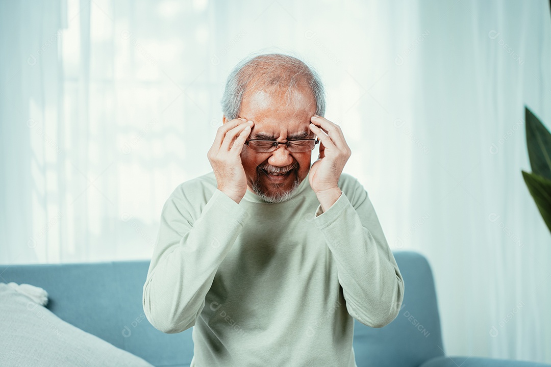 Idoso asiático sentindo dor de cabeça, sofrimento e enxaqueca na sala de estar em casa.
