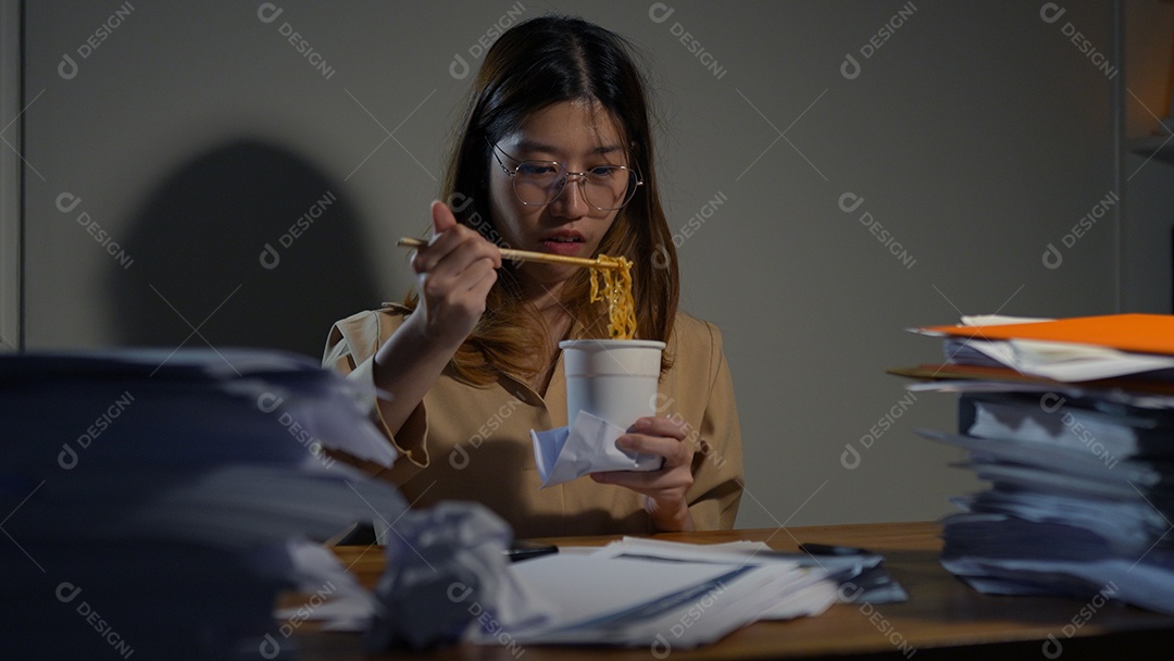 Empresária asiática comendo macarrão em copo de junk food entre trabalhar duro à noite