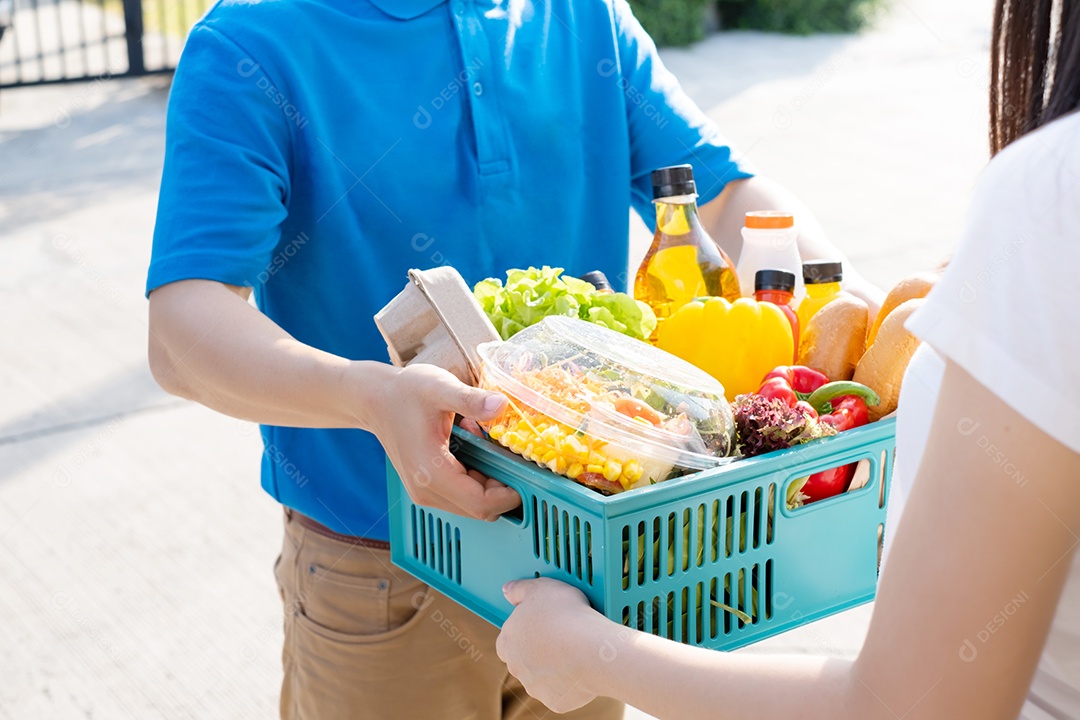 O homem asiático de entrega de comida em uniforme azul dá frutas e vegetais para a casa da frente do cliente recepto