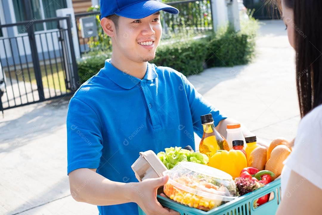 O homem asiático de entrega de comida em uniforme azul dá frutas e vegetais para a casa da frente do cliente recepto