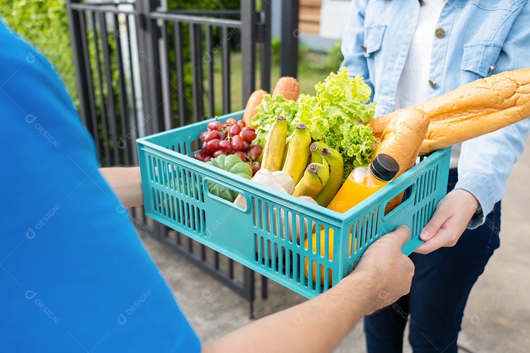 O homem asiático de entrega de comida em uniforme azul dá frutas e vegetais para a casa da frente do cliente receptor