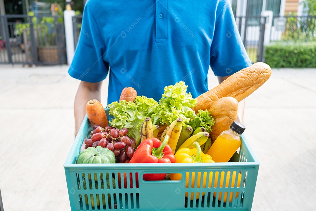 O homem asiático de entrega de comida em uniforme azul dá frutas e vegetais para a casa da frente do cliente receptor