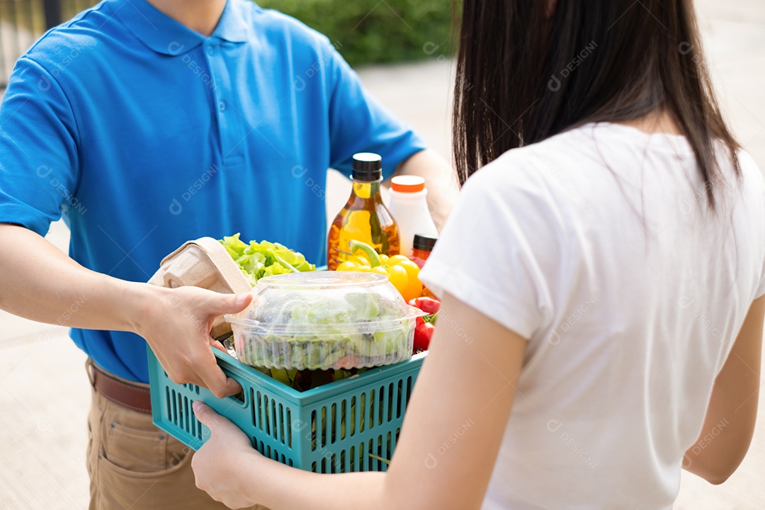 Homem asiático de entrega de comida em uniforme azul dá frutas e vegetais para a casa da frente do cliente receptor