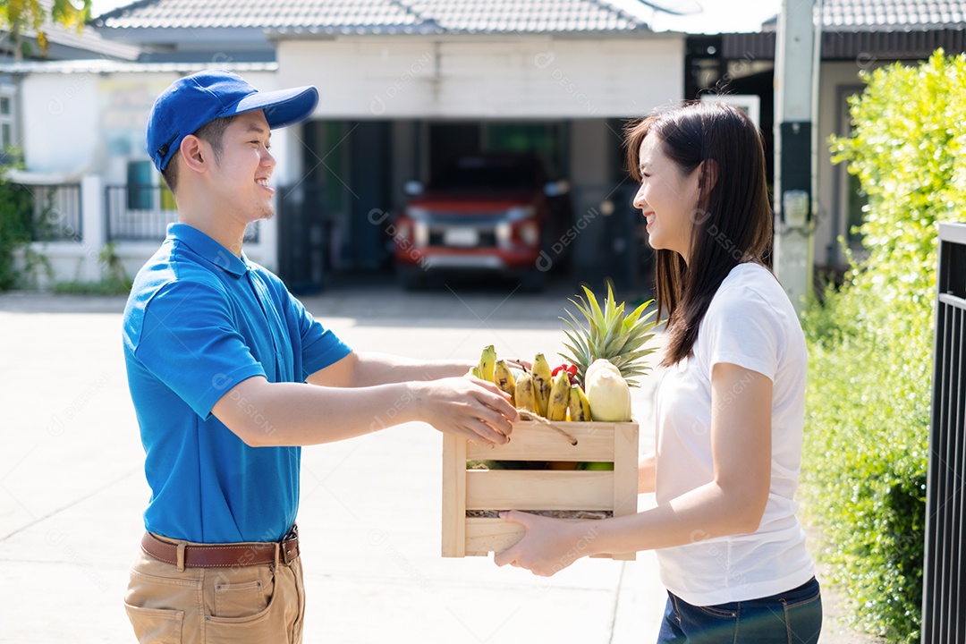 Homem asiático de entrega de comida em uniforme dá frutas e vegetais para a casa da frente do cliente receptor