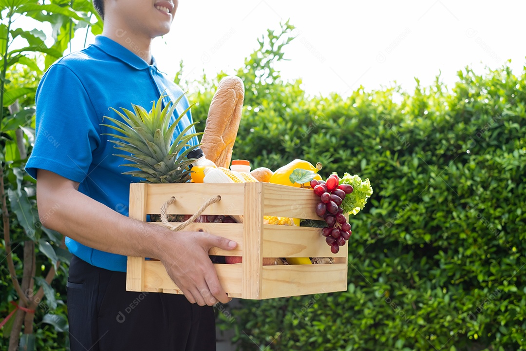 O homem asiático de entrega de comida em uniforme azul dá frutas e vegetais para a casa da frente do cliente receptor
