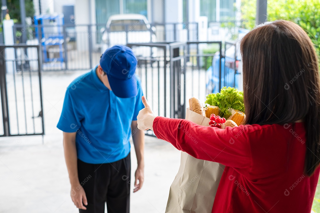 O entregador de comida usando máscara dá supermercado de compras de frutas e vegetais para a casa da frente do cliente