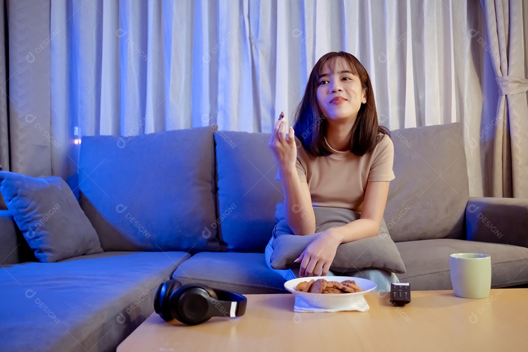 Jovem mulher asiática comendo sentada no sofá e assistindo televisão