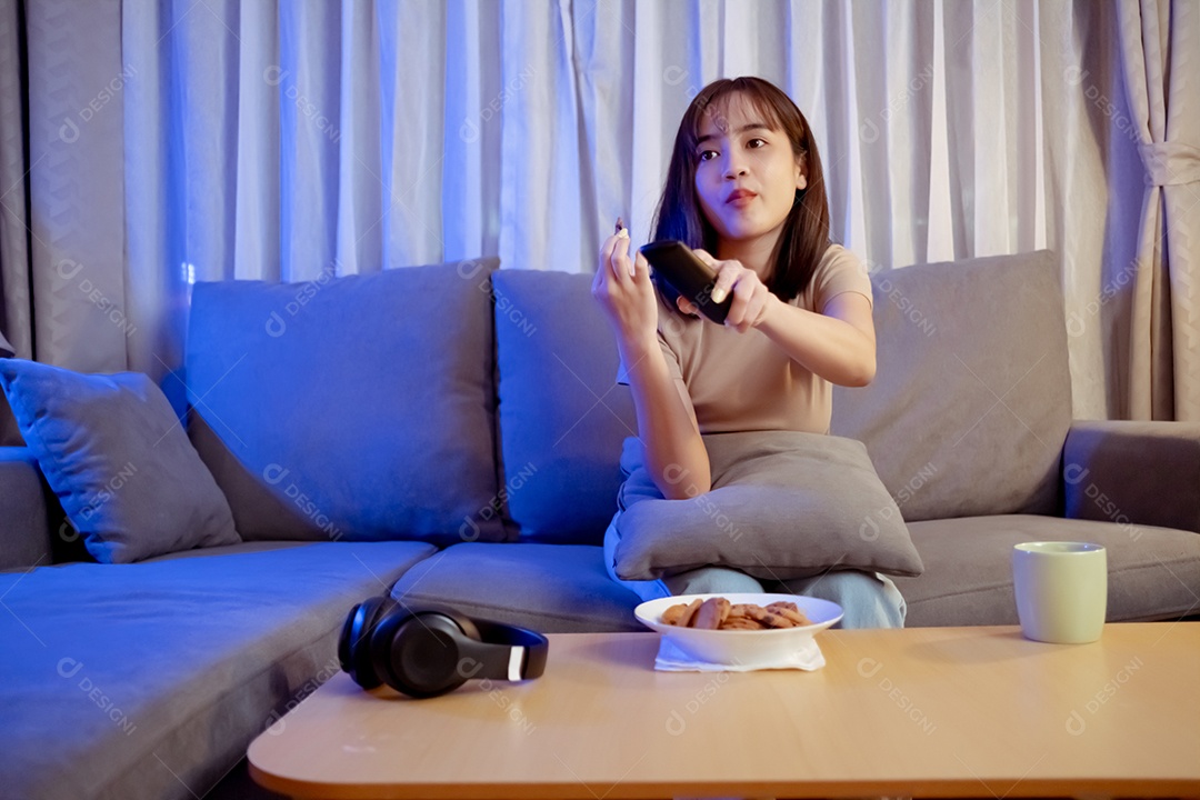 Jovem mulher asiática comendo sentada no sofá e assistindo televisão