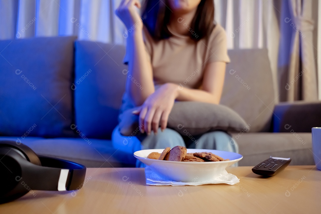 Jovem mulher asiática comendo sentada no sofá e assistindo televisão