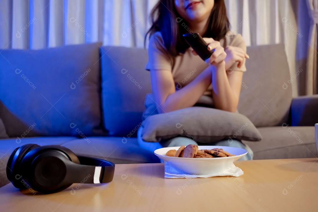 Jovem mulher asiática comendo sentada no sofá e assistindo televisão