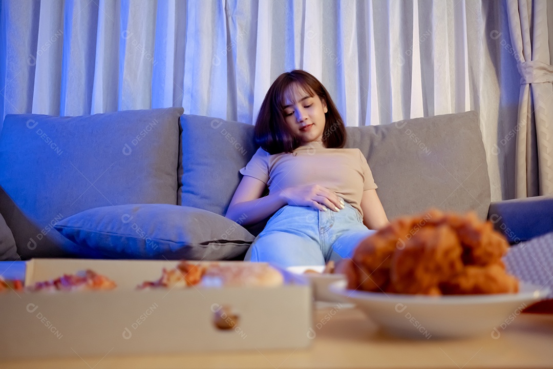 Jovem mulher asiática comendo sentada no sofá e assistindo televisão