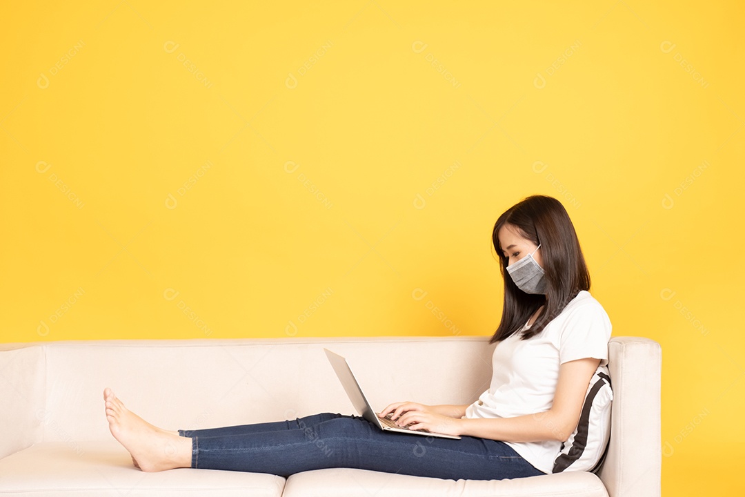 Jovem mulher asiática usando máscara facial sob o surto do vírus. Ela está sentada no sofá e fazendo uma chamada de vídeo