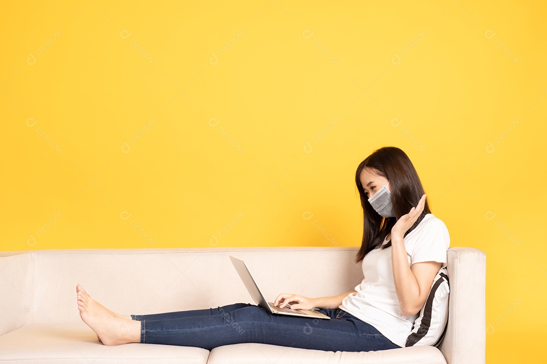 Jovem mulher asiática usando máscara facial sob o surto do vírus. Ela está sentada no sofá e fazendo uma chamada de vídeo