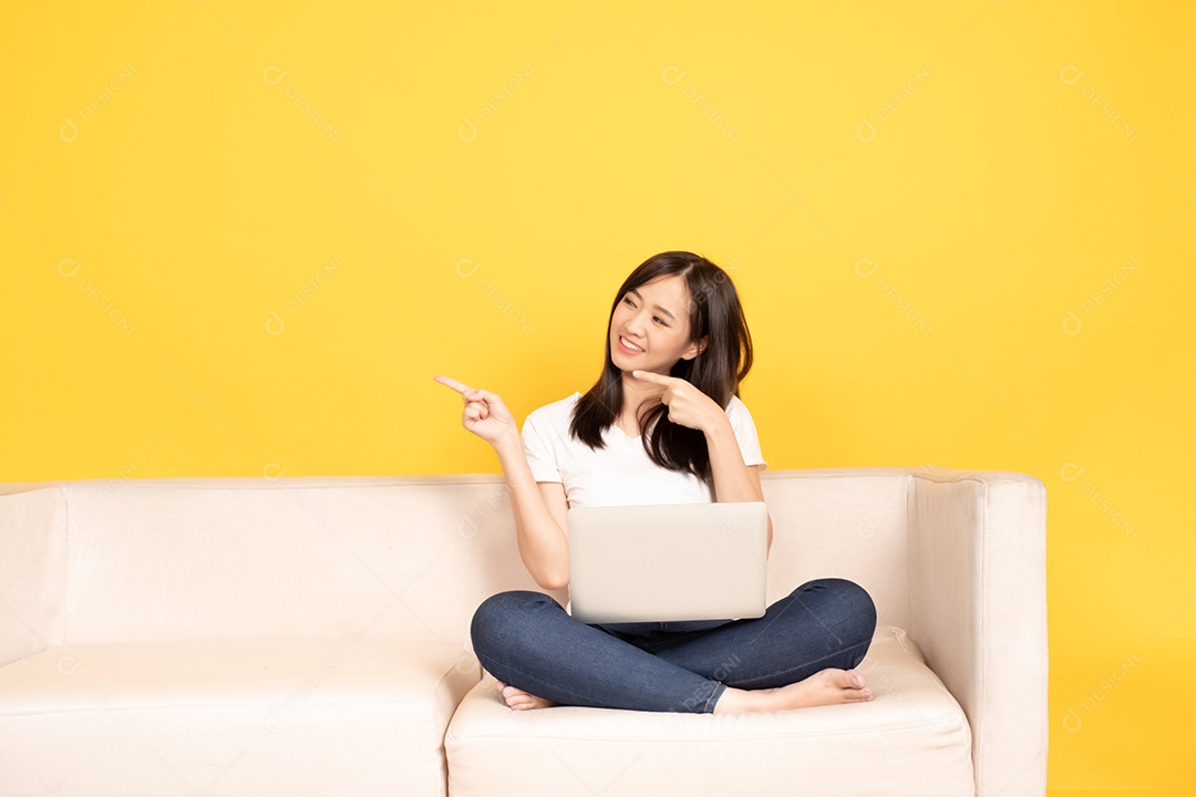 A jovem asiática aponta o dedo no sofá confortável, usando o laptop em casa.
