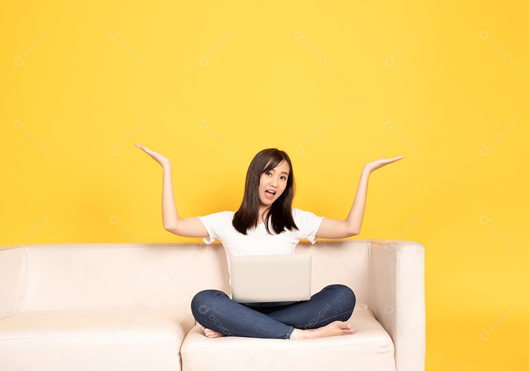 A jovem asiática aponta o dedo no sofá confortável, usando o laptop em casa.