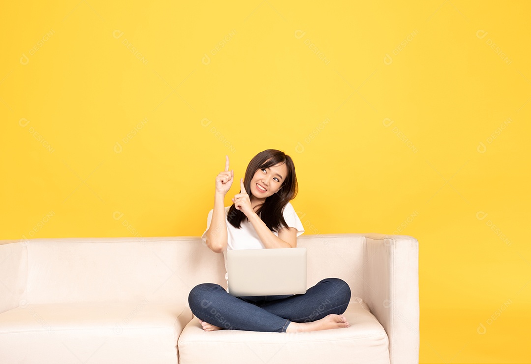A jovem asiática aponta o dedo no sofá confortável, usando o laptop em casa.