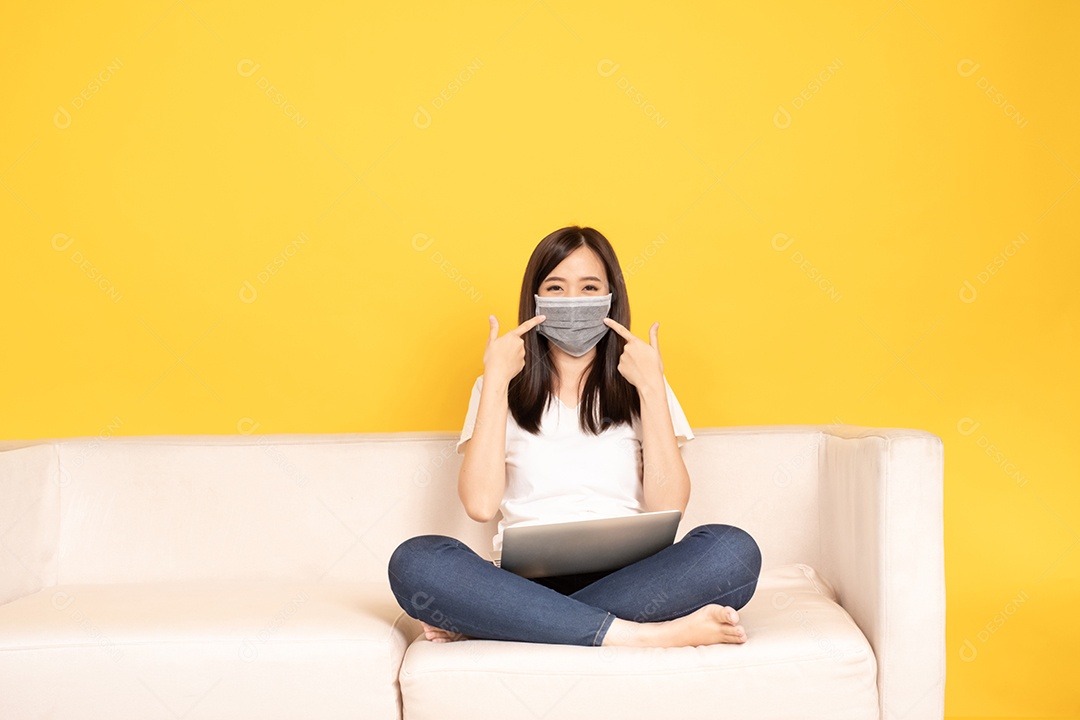 A jovem asiática aponta o dedo no sofá confortável, usando o laptop em casa.