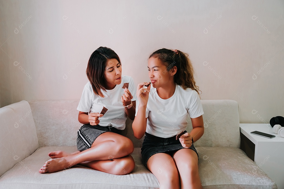 Mãe e filha adolescente comendo barra de chocolate no sofá