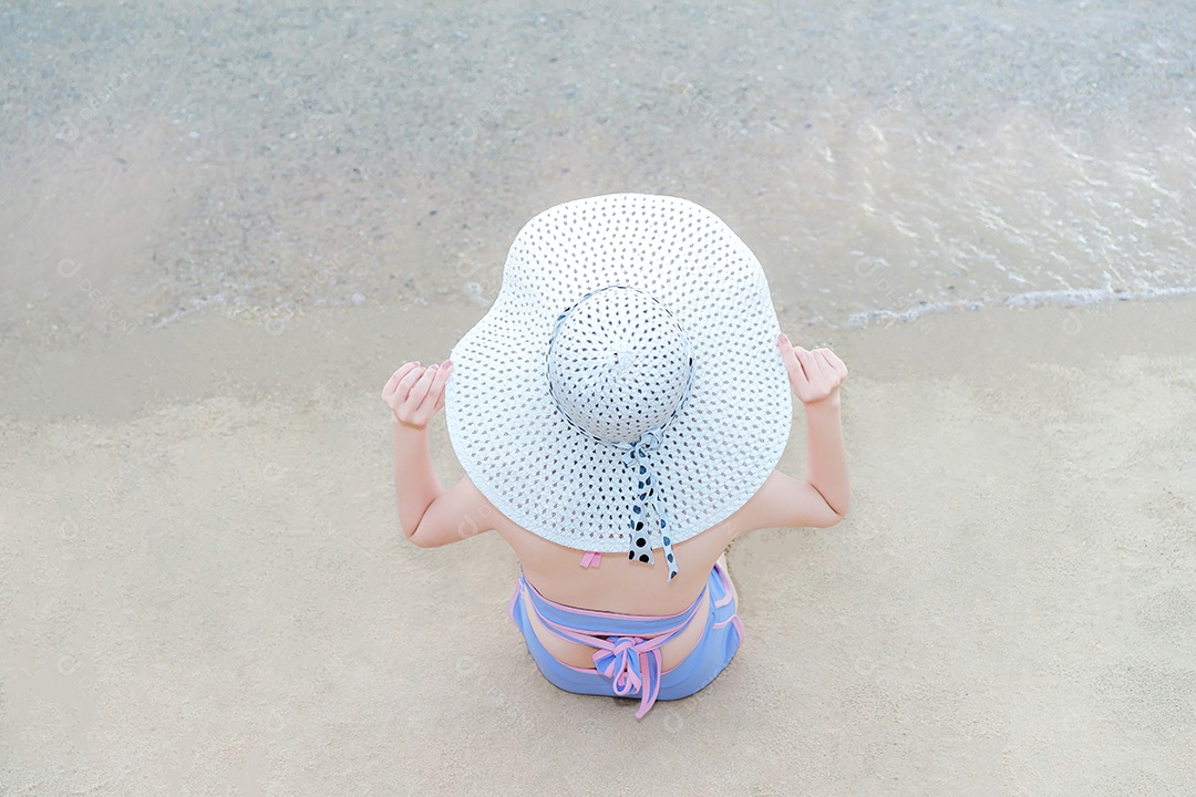 Jovem garota de biquíni com chapéu de havaí sentado na areia branca na praia à beira-mar