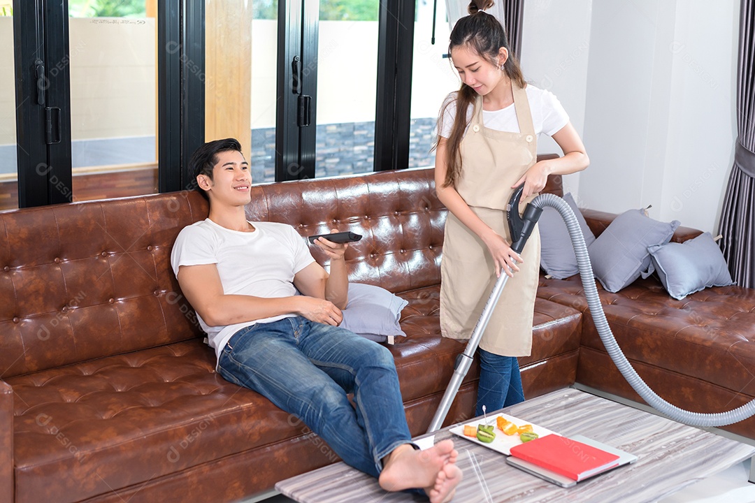 Casal limpando sala de estar em casa, jovem asiático segurando controle remoto assistindo tela de televisão