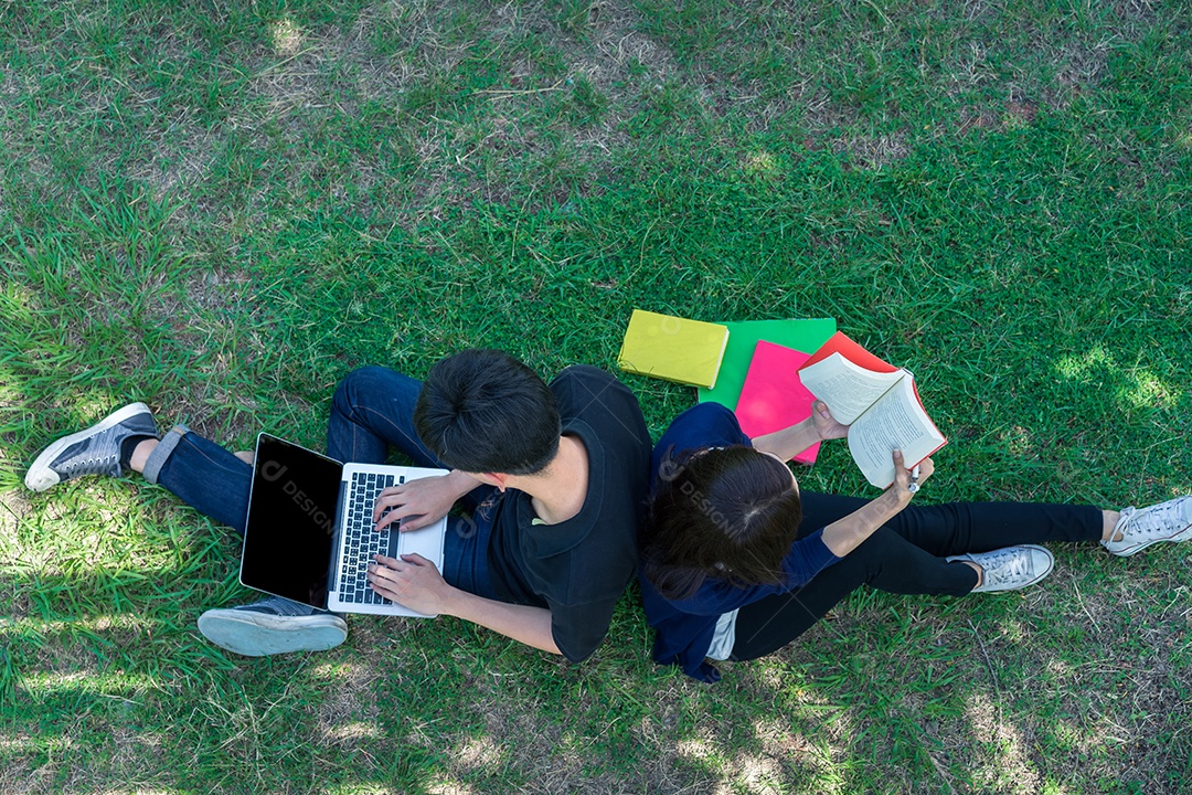 Jovens estudantes juntos sentados na grama abrem pastas escolares e usando computador portátil