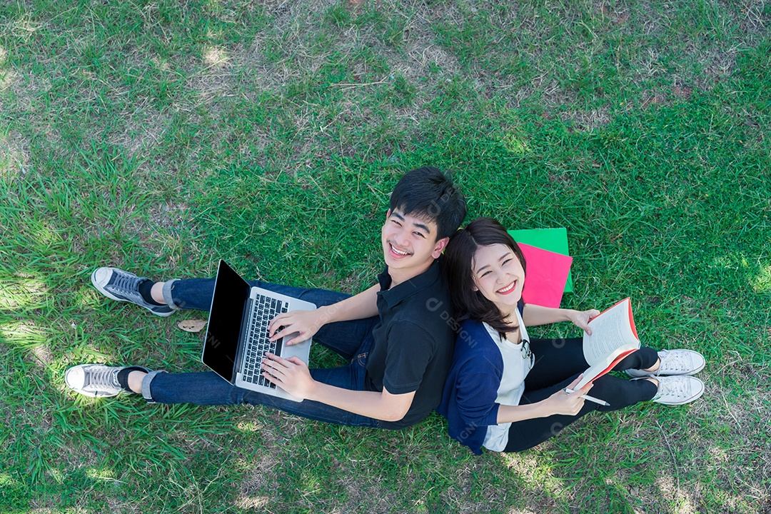 Jovens estudantes juntos sentados na grama abrem pastas escolares e usando computador portátil