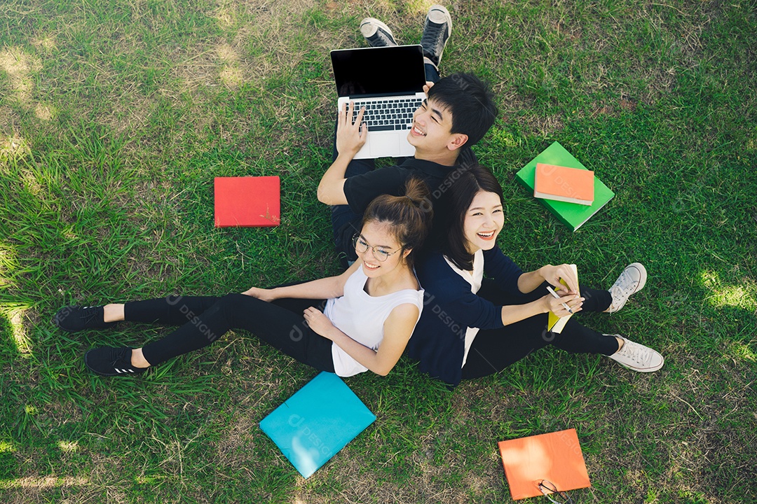 Jovens estudantes juntos sentados na grama abrem pastas escolares e usando computador portátil