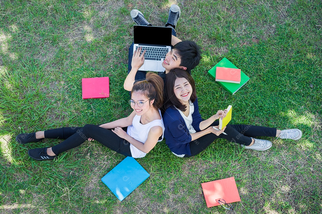 Jovens estudantes juntos sentados na grama abrem pastas escolares e usando computador portátil