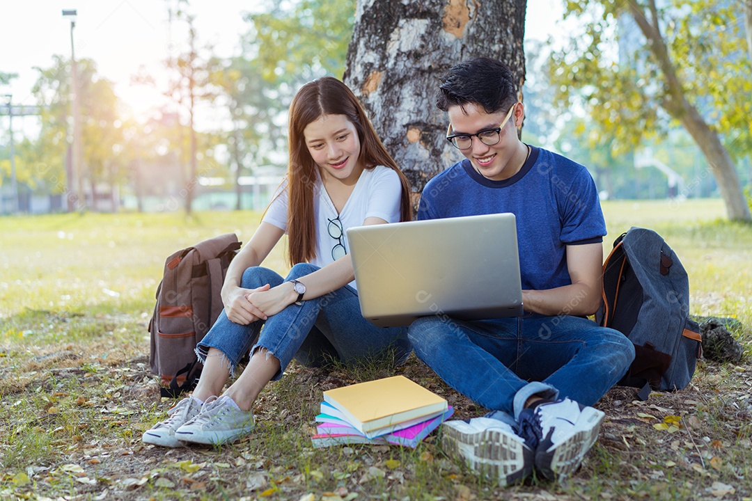 Estudantes jovens asiáticos juntos usando consulta de computador portátil e lendo livro