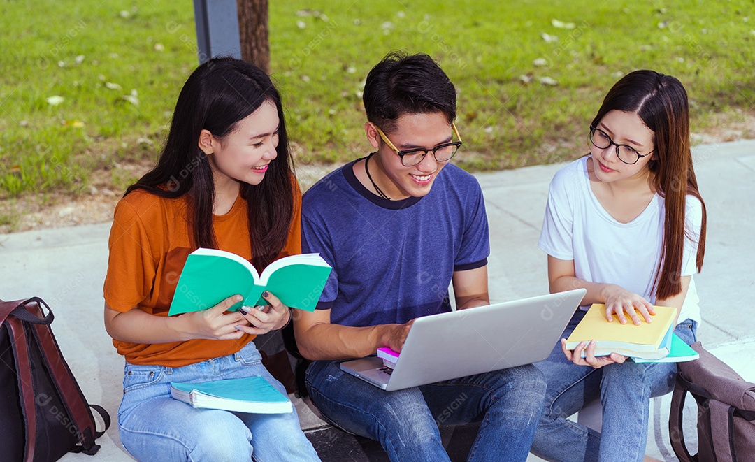 Estudantes jovens asiáticos juntos usando consulta de computador portátil e lendo livro