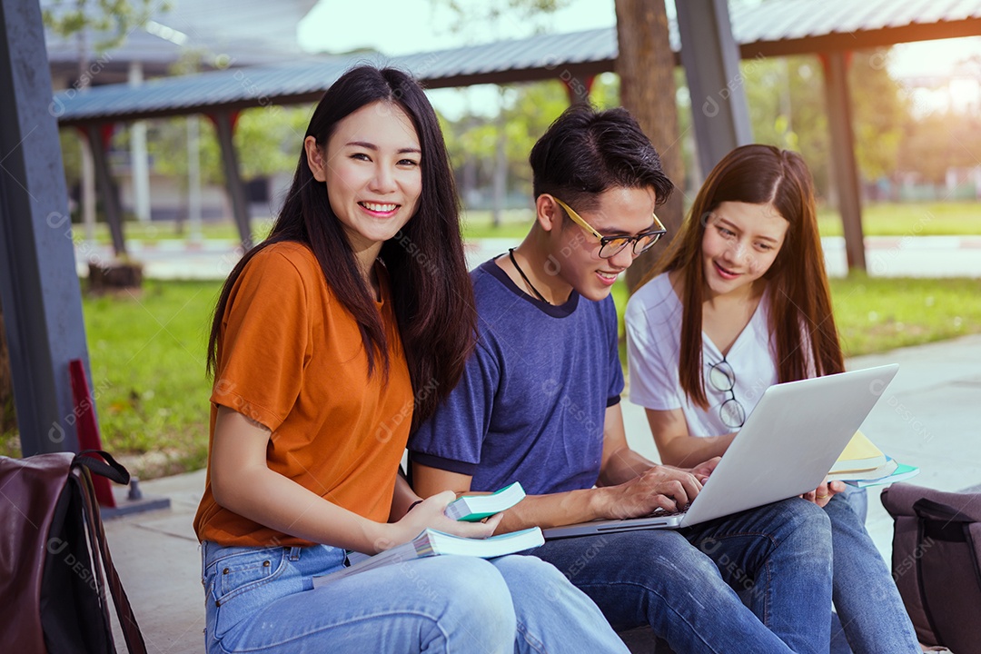Estudantes jovens asiáticos juntos usando consulta de computador portátil e lendo livro
