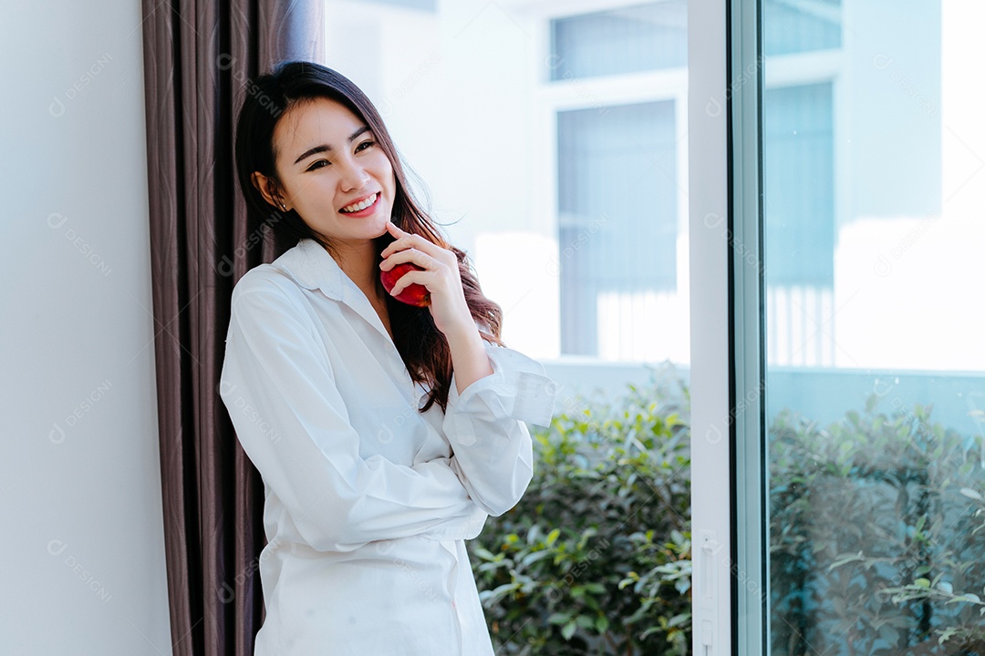 Mulher asiática segurando maçã perto da janela na sala de estar