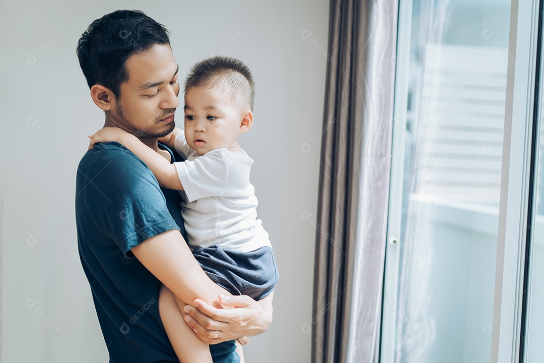 Pai asiático segura filho pequeno perto da janela em casa.