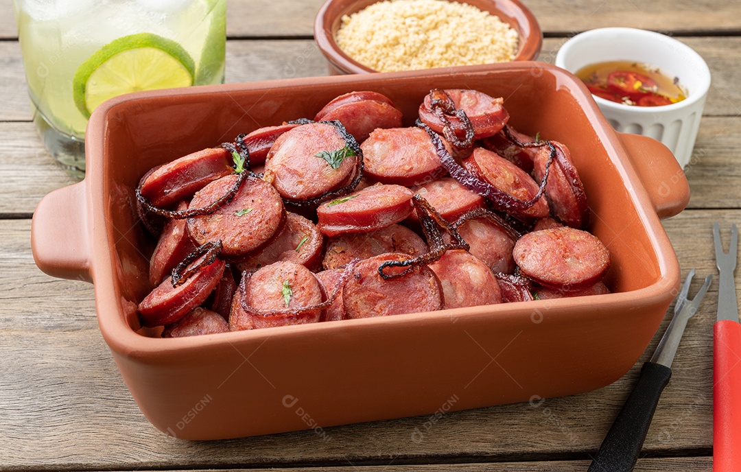 Porção de linguiça calabresa grelhada com cebola, farofa e caipirinha sobre mesa de madeira.