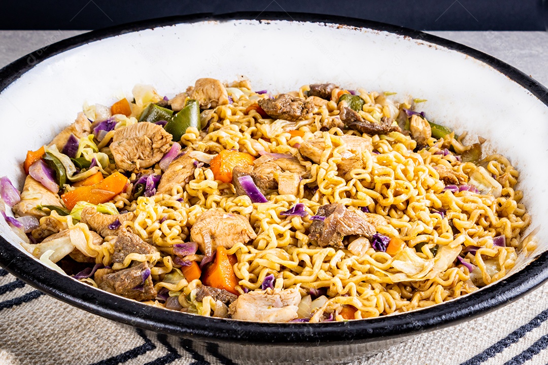 Yakisoba japonês com macarrão frito em panela metálica branca na mesa com toalha de mesa branca e fundo preto, com pauzinhos de comida japonesa... Close view.